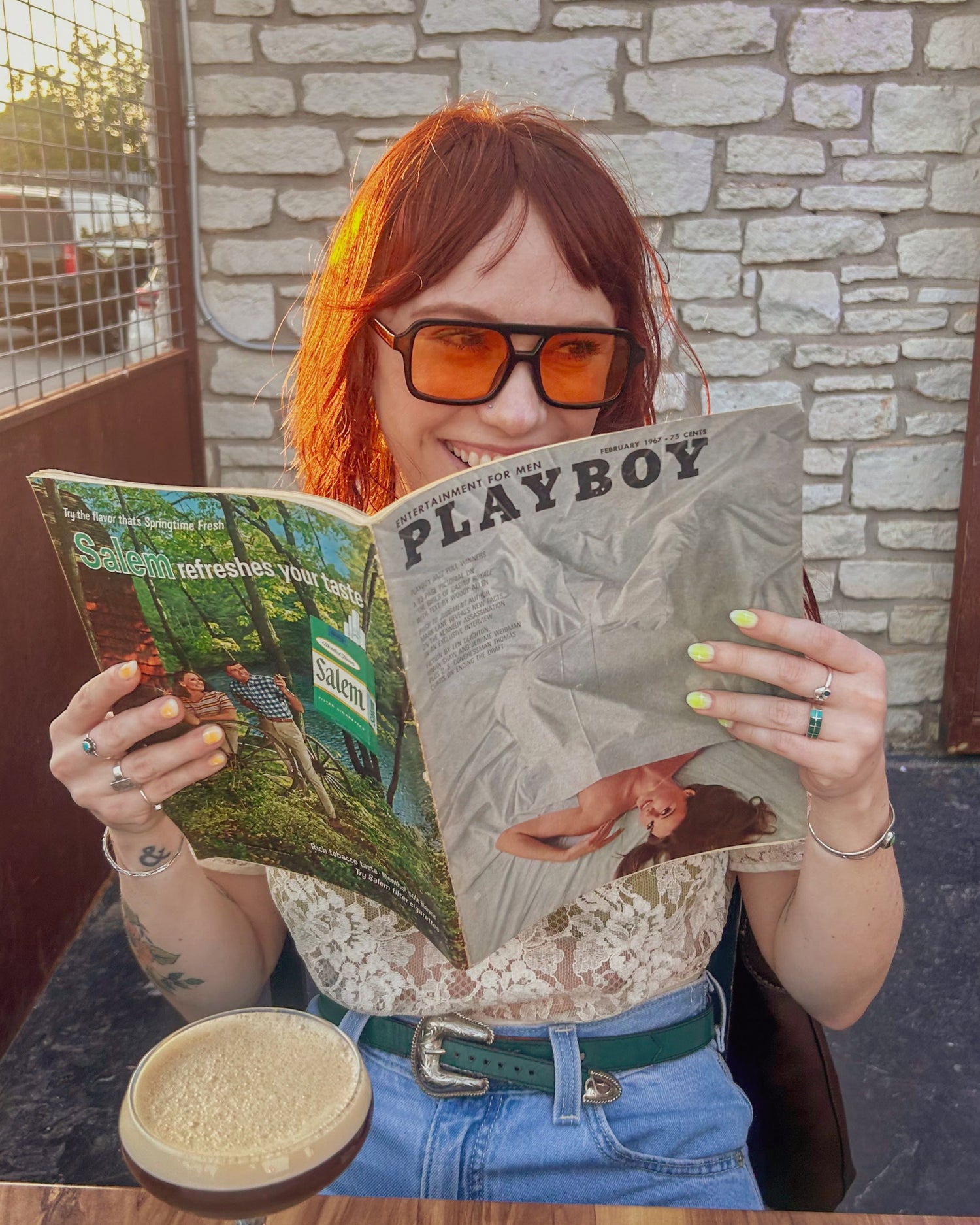 Person with red hair and orange glasses reading a Playboy magazine outdoors.