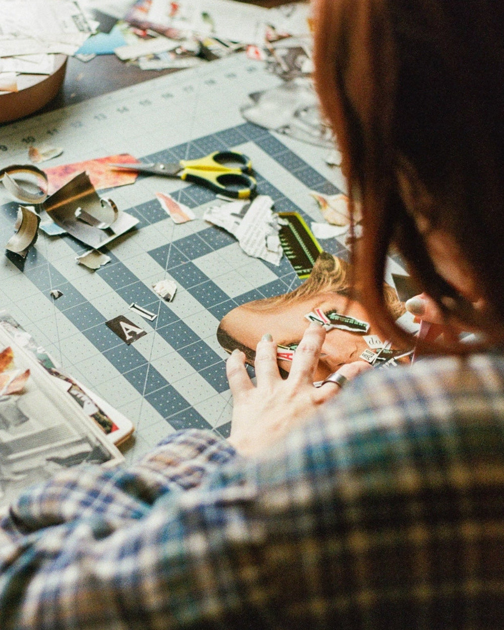 A person with long red hair, wearing a blue and green plaid shirt, is seen from behind looking down at a craft table cluttered with paper, scissors, and tools.
