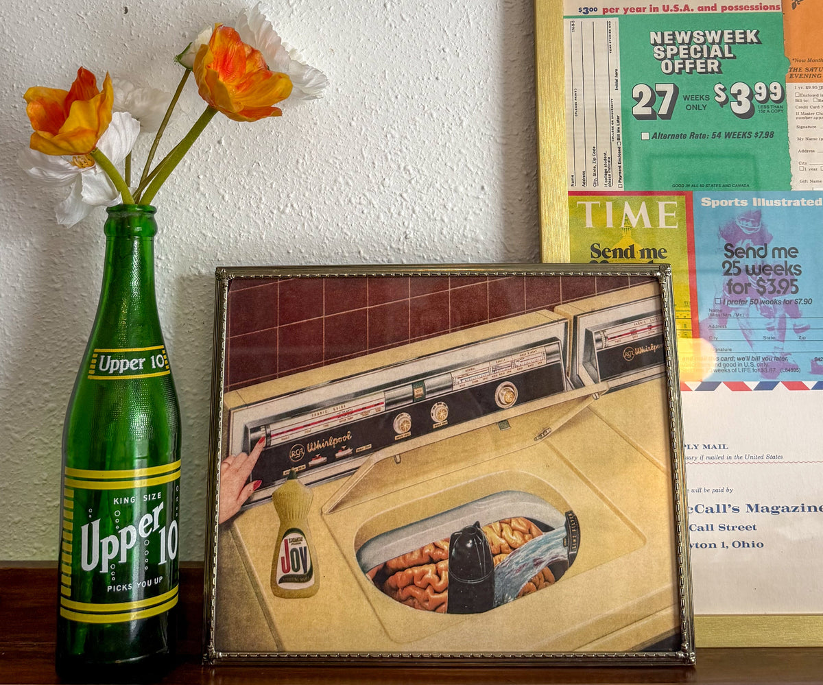 A framed vintage-style print showing a retro washing machine scene, displayed on a wooden surface next to a green Upper 10 bottle vase with orange and white flowers, against a white wall with old posters behind it.