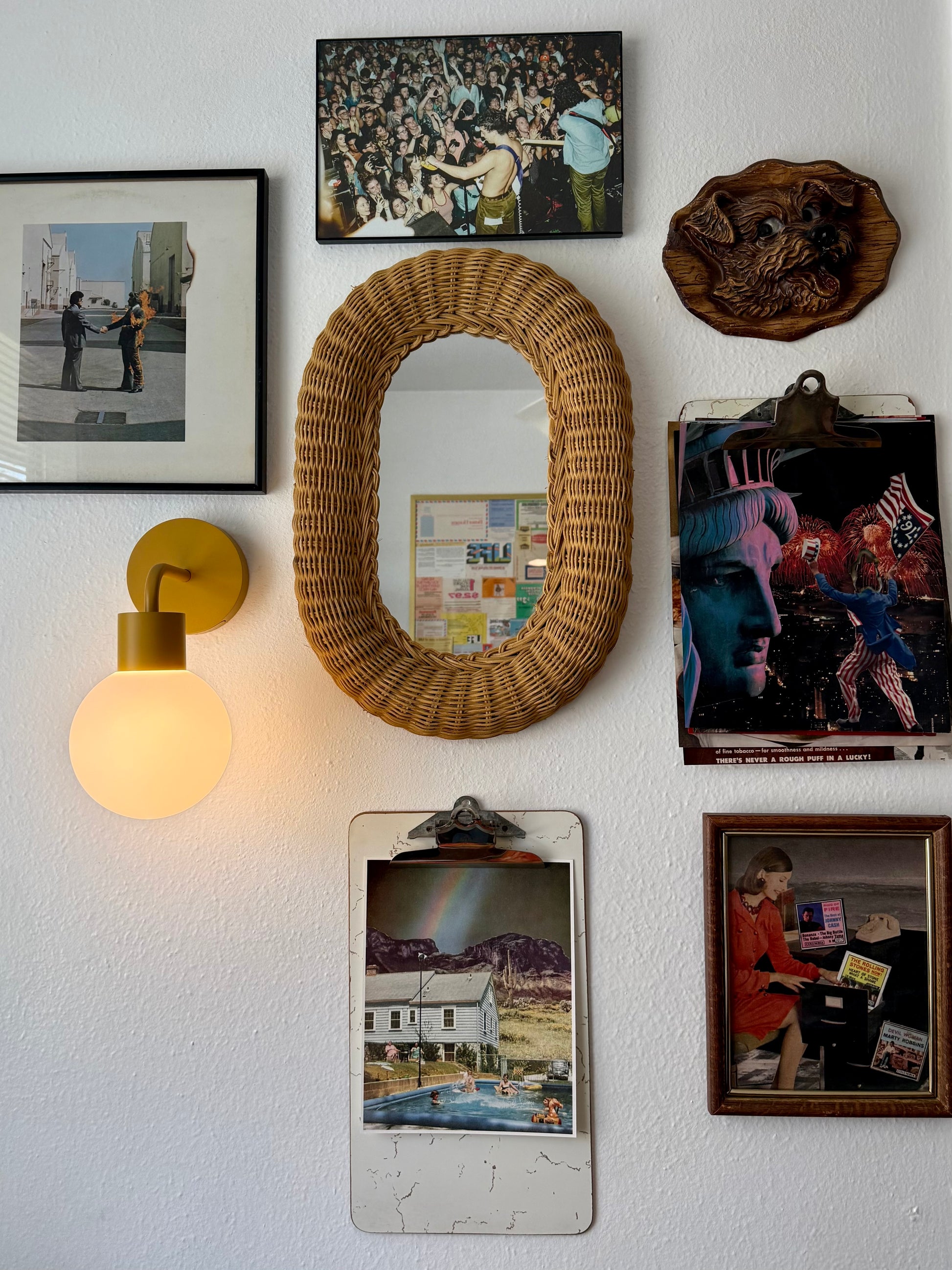 A styled gallery wall featuring a large oval wicker mirror, a glowing yellow wall sconce, multiple framed and clipped photographs, and a carved wooden plaque on a textured white wall.