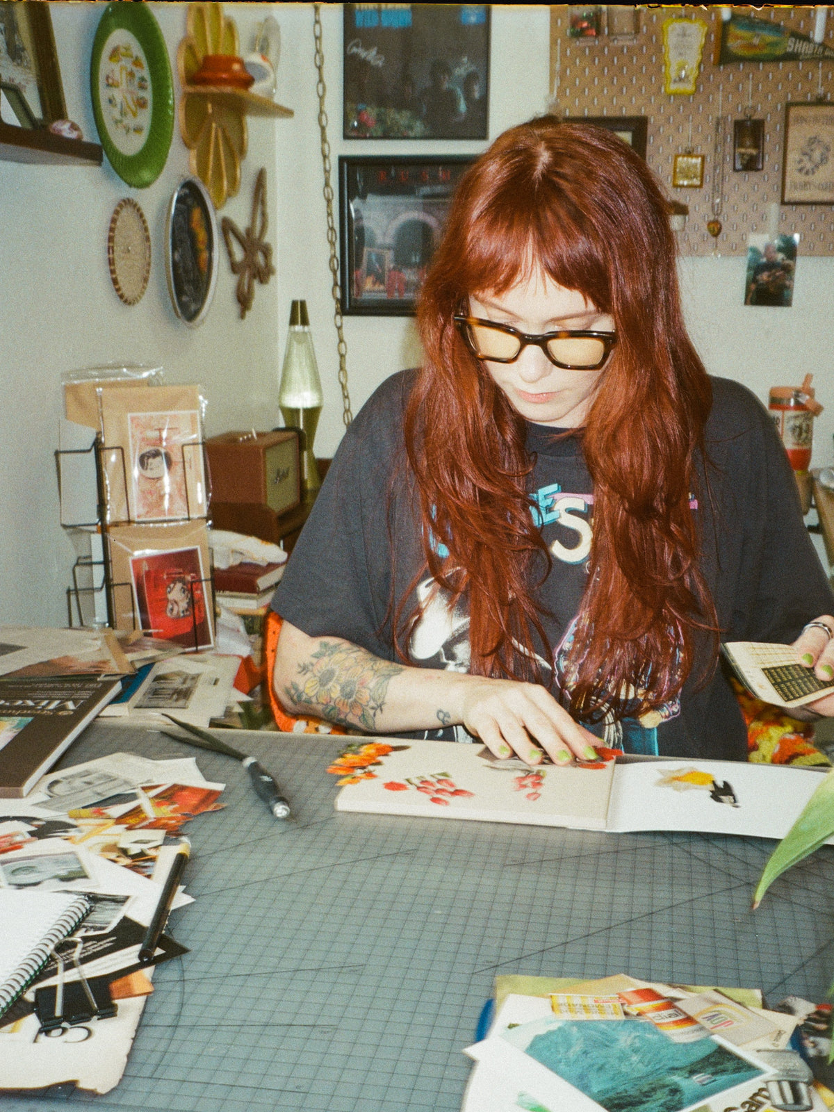 A woman with long red hair and glasses sits at a cluttered desk, working on a collage. The room has a vintage, creative studio feel with shelves, decorative plates, and papers scattered across the table.