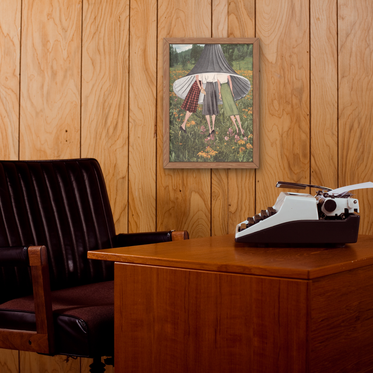 A framed art print depicting three women standing in a field, displayed on a wood-paneled wall above a wooden desk with a vintage typewriter and a dark leather office chair.