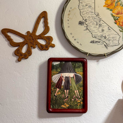 A framed art print featuring an illustration of three women in a field under a UFO, displayed on a textured white wall alongside a wooden butterfly ornament and a decorative plate.