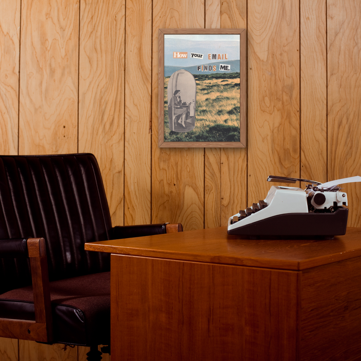 A framed art print featuring a woman at a desk with the text 'HOW YOUR EMAIL FINDS ME' displayed on a wood-paneled wall above a wooden desk with a typewriter and a dark leather office chair.