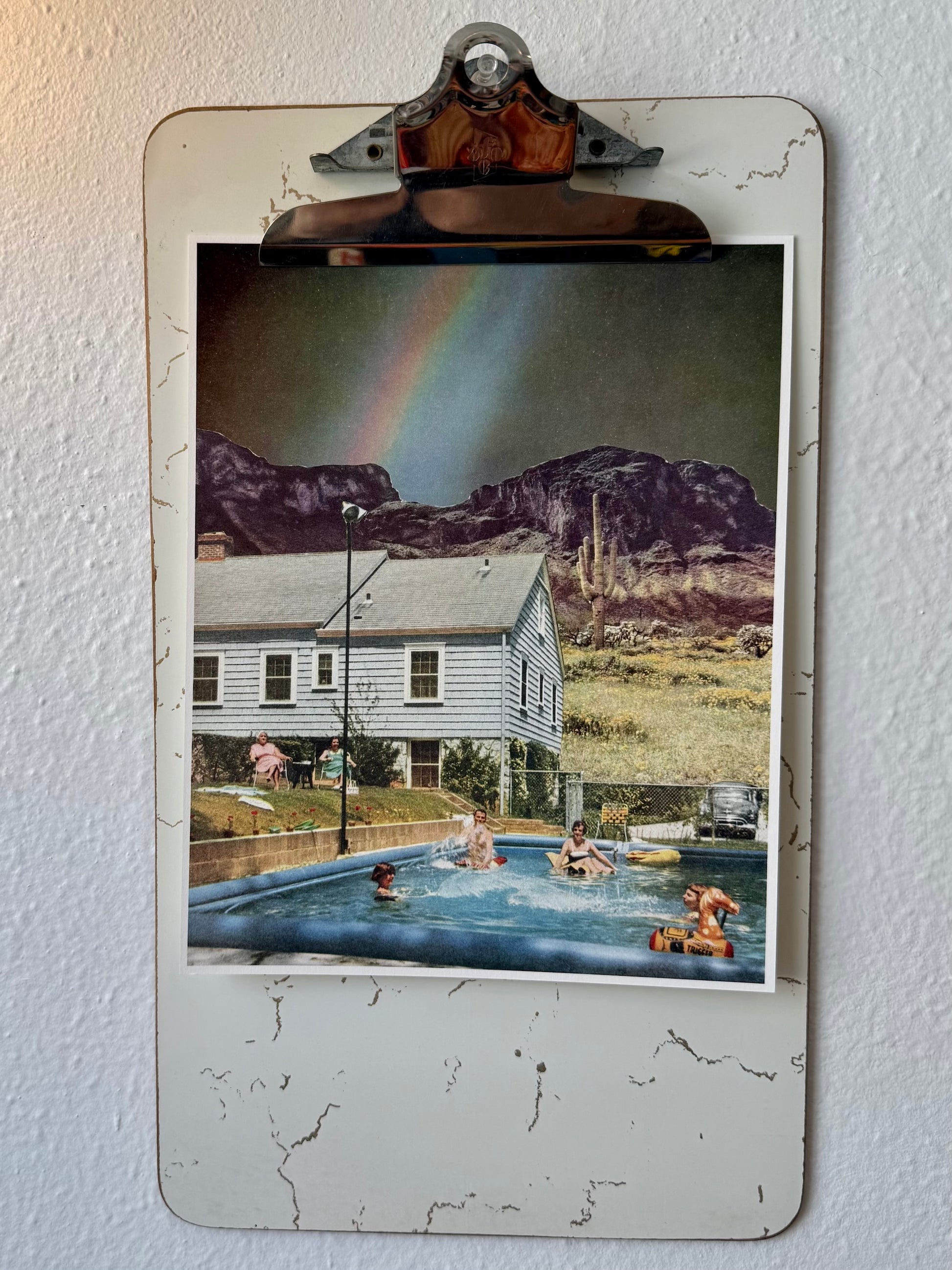 A vintage-style distressed metal clipboard with a binder clip, mounted on a textured white wall. The clipboard holds a square printed photograph depicting a poolside scene with people, a house, mountains, and a rainbow.