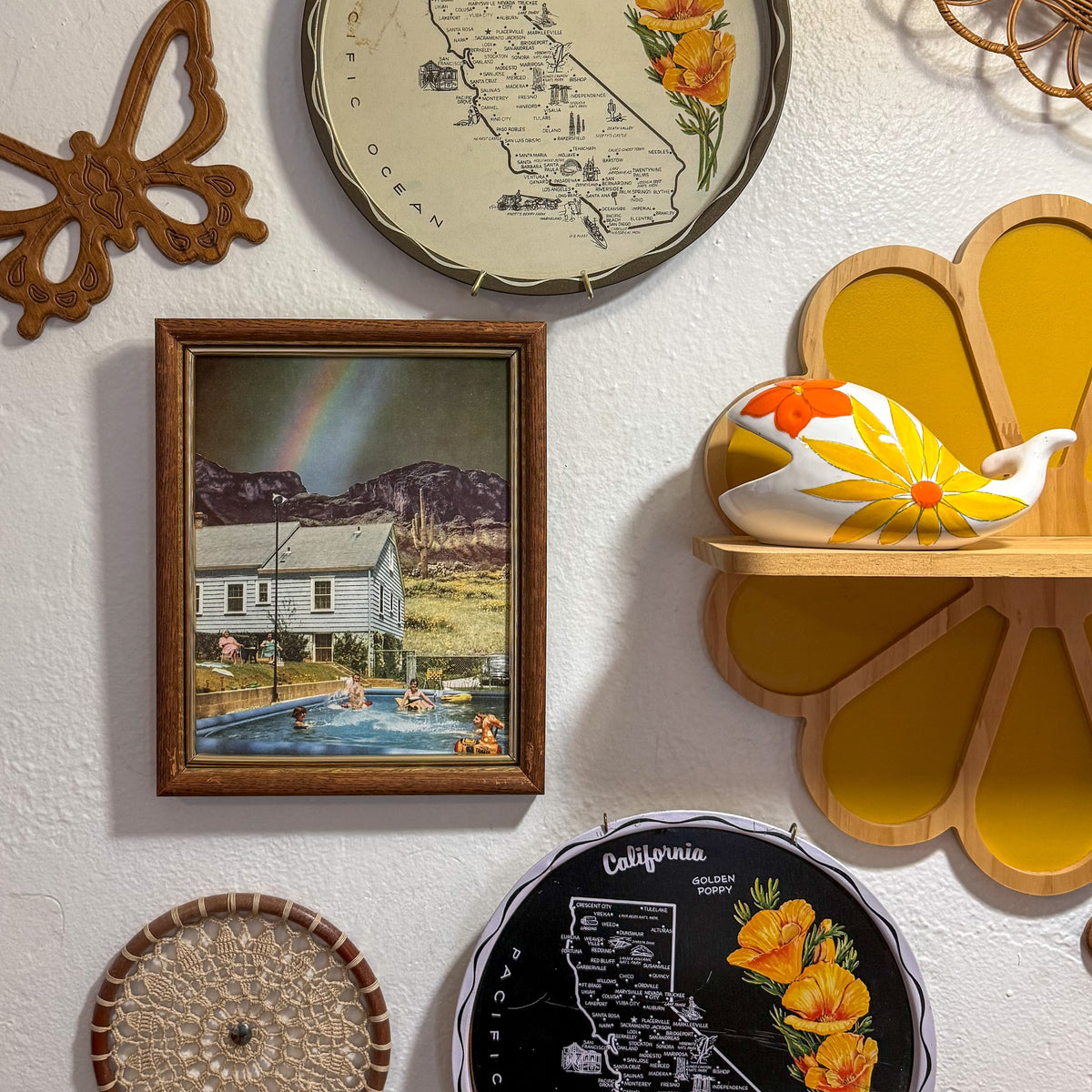 A styled wall display featuring a framed vintage-style painting of a house and pool, a California map print with flowers, a black California map sign, a butterfly wall decor piece, a wooden shelf with a floral ceramic pitcher, and other decorative items on a textured white wall.