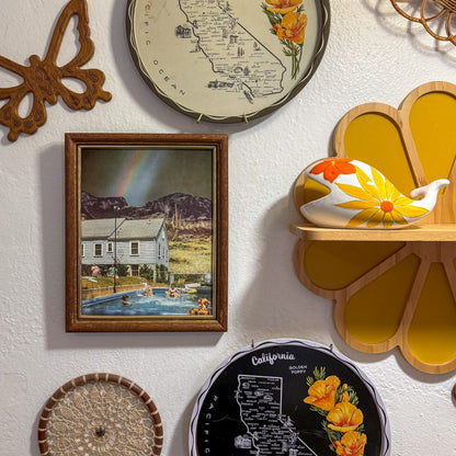 A styled wall display featuring a framed vintage-style painting of a house and pool, a California map print with flowers, a black California map sign, a butterfly wall decor piece, a wooden shelf with a floral ceramic pitcher, and other decorative items on a textured white wall.